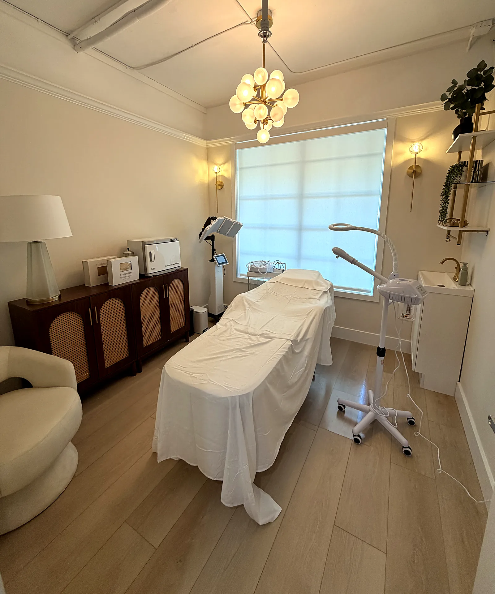 Treatment room at Skin Loft Studio: a draped facial bed beneath a brass chandelier, with a window and a cane cabinet.